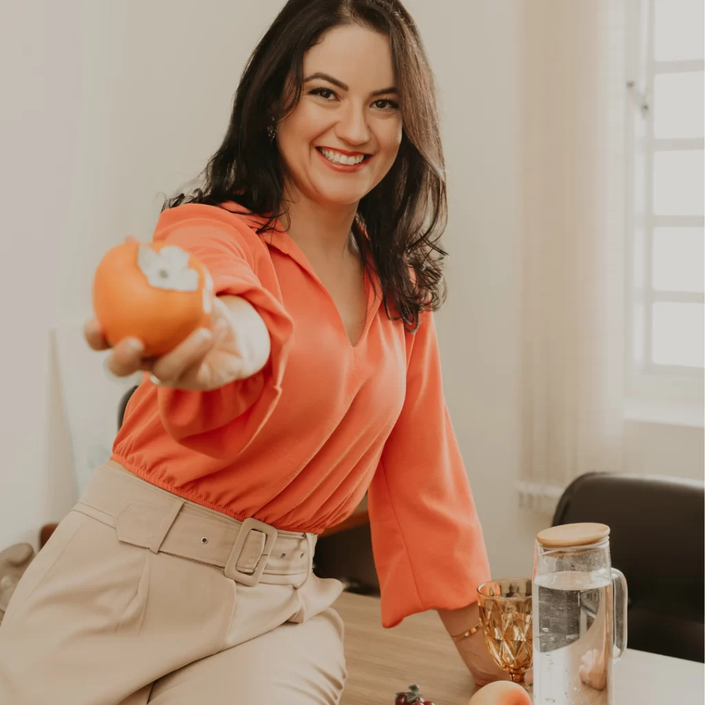 Mariana Drago sorrindo, estendendo um caqui para a câmera